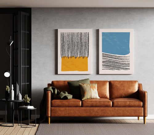A modern living room scene featuring a brown leather sofa with decorative pillows, two framed abstract wall art prints hanging above it, and a gray textured wall behind. The left print has black vertical stripes on a yellow background, and the right print has black horizontal lines on a blue and white background.